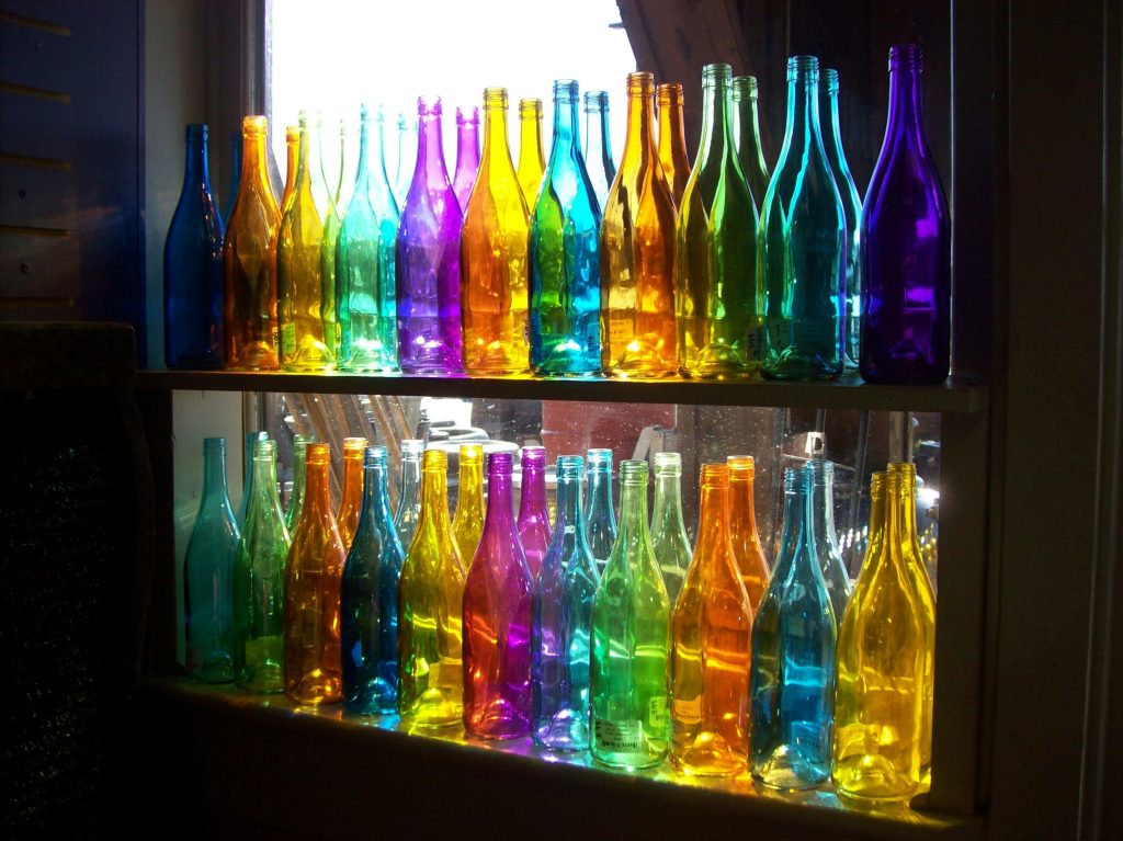 Two shelves of coloured glass bottles witha. mirror behind them in a dark room