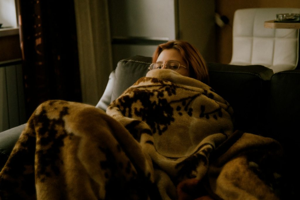 A woman wrapped in a heavy blanket on a sofa, only her eyes and glasses showing